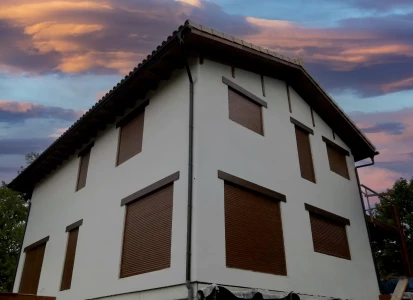 Aplicación de sistema SATE y acabado en corcho proyectado de textura ELASTICOVER FINE. Casa unifamiliar de dos plantas con la fachada de color blanco y muchas ventanas con las persianas bajadas de dolor marrón oscuro, a la que se ha aplicado el sistema de aislamiento térmico y acabado con ELASTICOVER FINE.