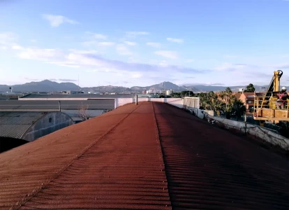 Aplicación de corcho proyectado Elasticover Tech. Aplicación de corcho proyectado rojo, Elasticover Tech, a una cubierta curva de una nave. con otras naves y chalets alrededor.