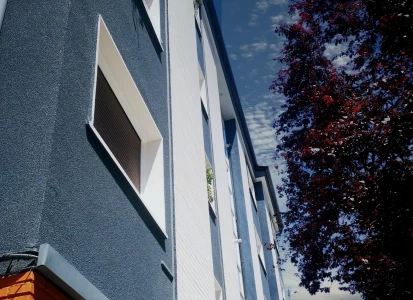 Aplicación de corcho proyectado ELASTICOVER TECH, en un edificio de viviendas. Vista hacia el cielo de la fachada de un edificio, con cuatro plantas de viviendas, con corcho proyectado ELASTICOVER TECH, en franjas verticales blancas y azules oscuras y un árbol a su lado